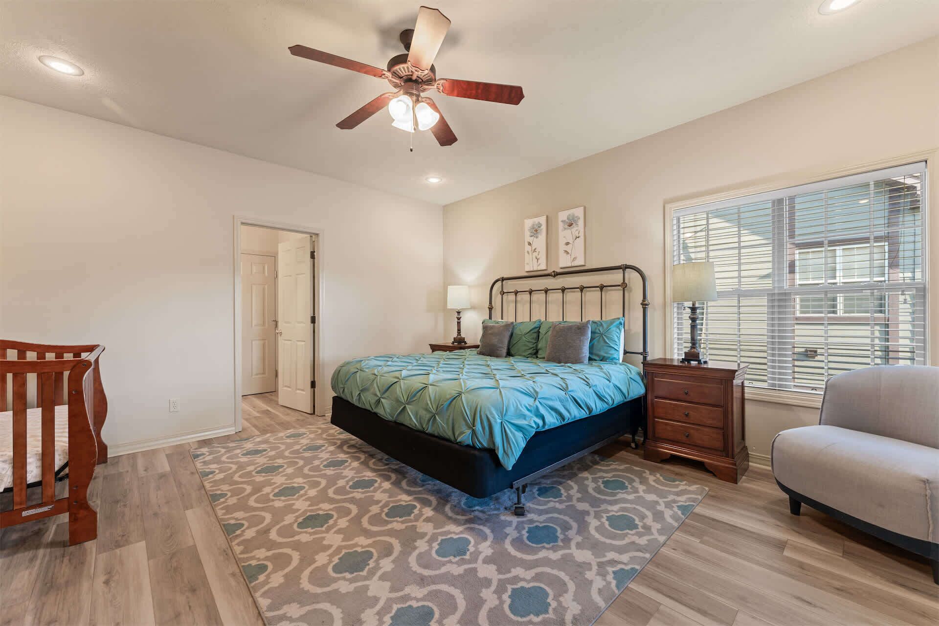 Quarto com cama, mesa de cabeceira, cadeira, banco de madeira, ventilador de teto, janela com persianas e porta.
