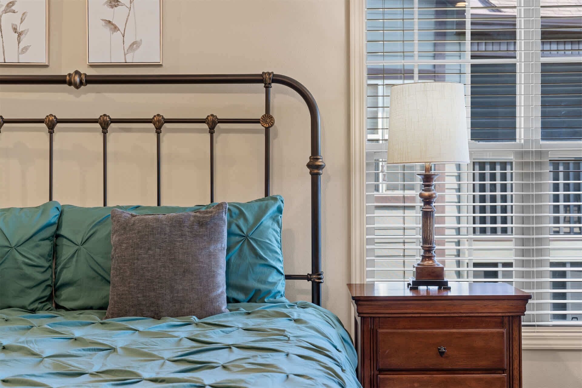 Un dormitorio con cama, almohadas, mesita de noche, lámpara y ventana con persianas.
