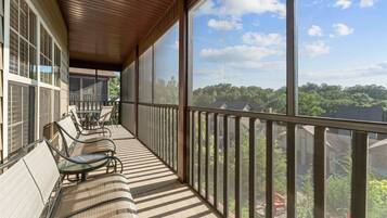 A covered balcony with a view of a forested area, a table with chairs, and a railing.