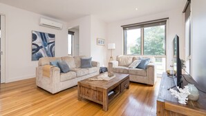 A spacious living room with two beige sofas, a wooden coffee table, and a large window providing a view of the outdoors. - Freda Beach Haven, centrally located (Inverloch)
