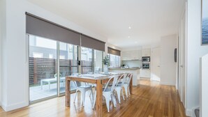 A modern kitchen with white cabinets, a dining area with a wooden table and white chairs, and a sliding glass door leading to a patio. - Freda Beach Haven, centrally located (Inverloch)