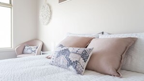 A neatly made bed with patterned pillows, a chair, and a hanging light fixture in a bright room with a window. - Freda Beach Haven, centrally located (Inverloch)