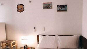 A hotel room with a neatly made bed, a wooden side table with a lamp, and wall decorations including a motorcycle poster and a sign.