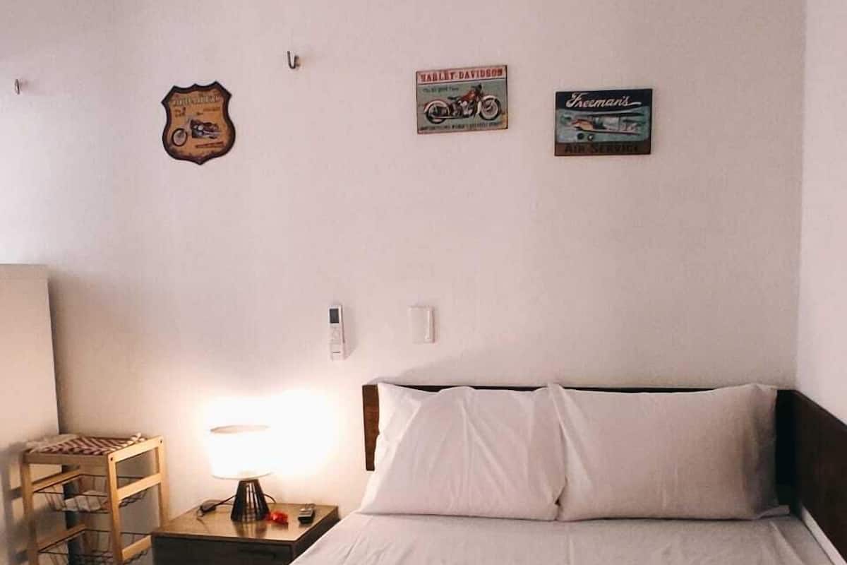 Ein Hotelzimmer mit einem ordentlich bezogenen Bett, einem Nachttisch aus Holz mit einer Lampe und Wanddekorationen, darunter ein Motorradplakat und ein Schild.