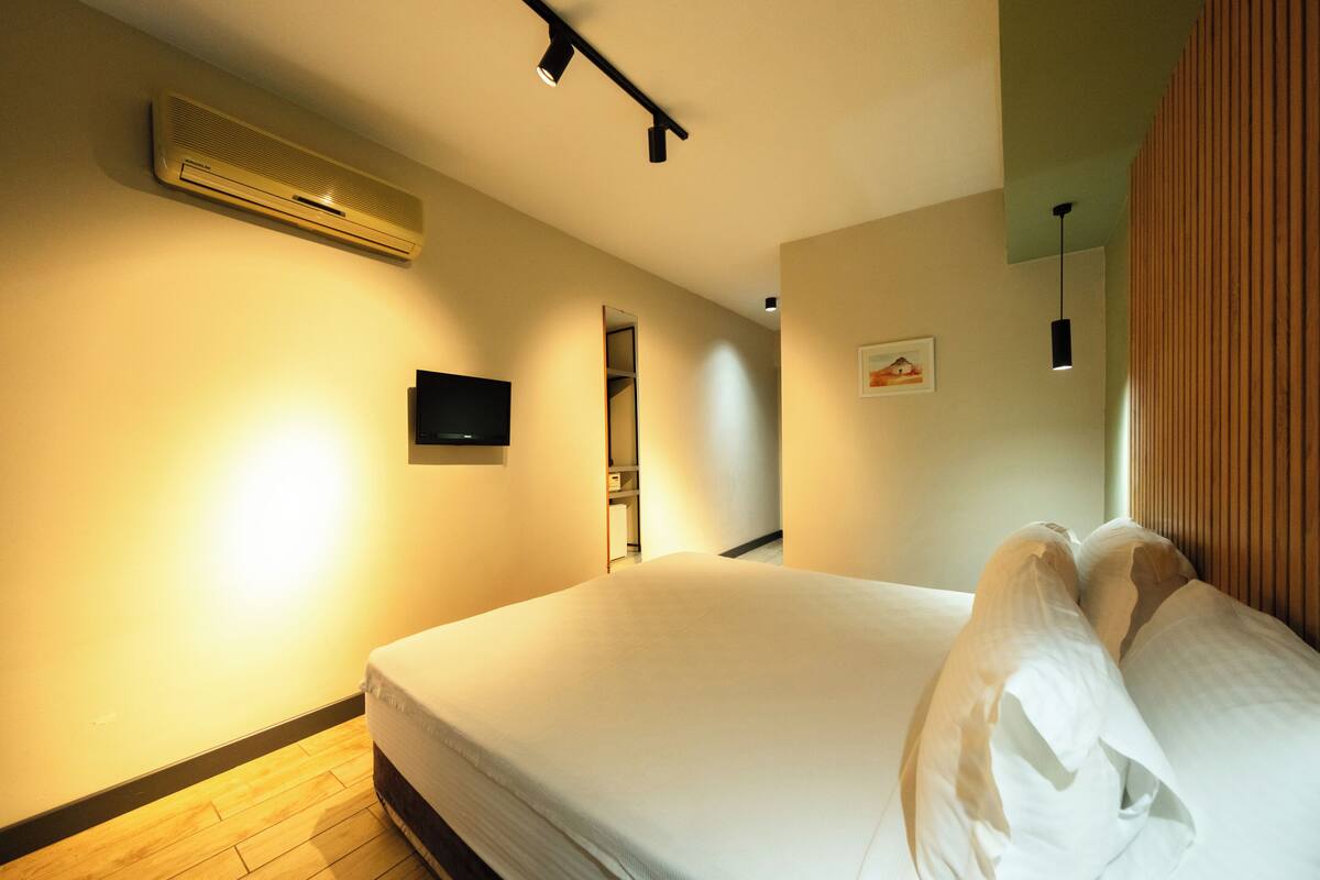 A hotel room with a bed, an air conditioner, a TV, and a wooden floor.