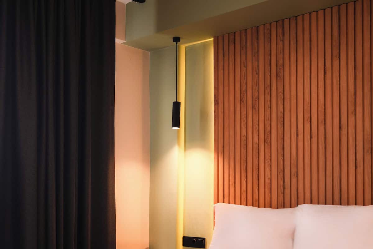 A modern hotel room with a large bed, dark curtains, wooden paneling on the wall, and a small bedside table with a lamp.