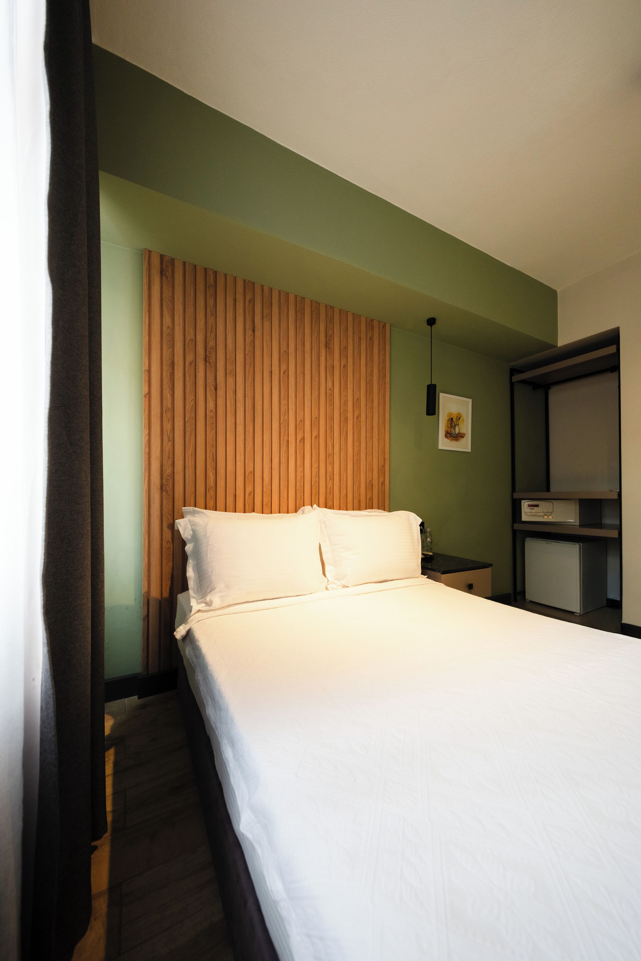 A hotel room with a bed, a wooden headboard, a nightstand, a small desk, and a shelf with a picture.