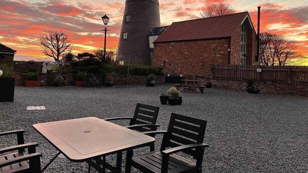 Un edificio rústico con fachada de ladrillo y tejado de tejas rojas, una alta torre cilíndrica y un patio de grava con mesas al aire libre.