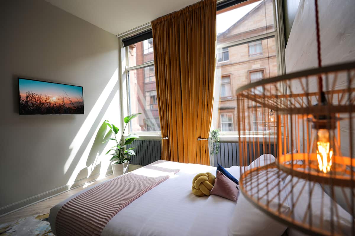 Ein Hotelzimmer mit Bett, Zimmerpflanze, Wandfernseher und einem Fenster mit Vorhängen.