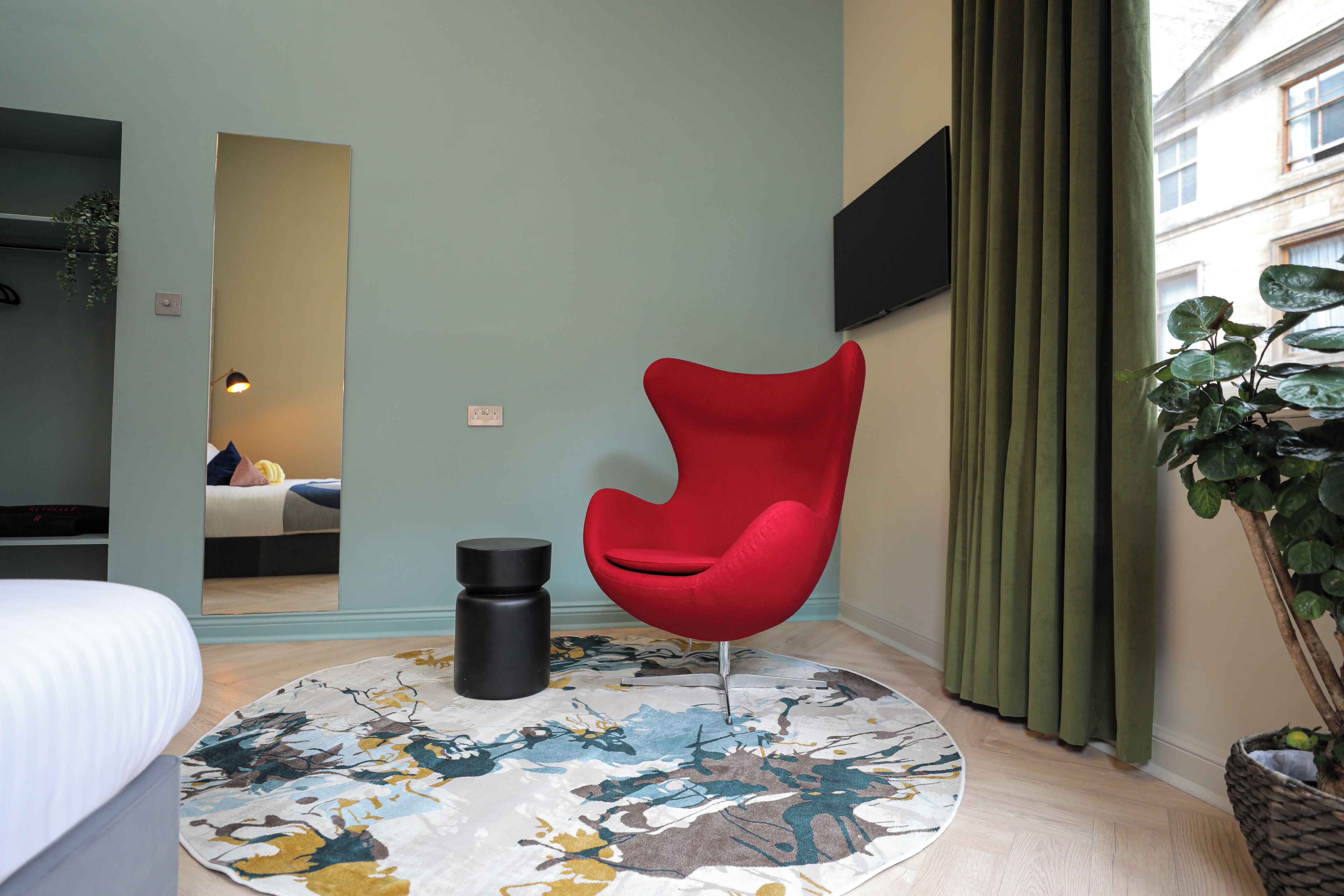 Habitación de hotel moderna con una silla roja, un tapete redondo con estampado floral, un cesto de basura negro y un espejo grande.