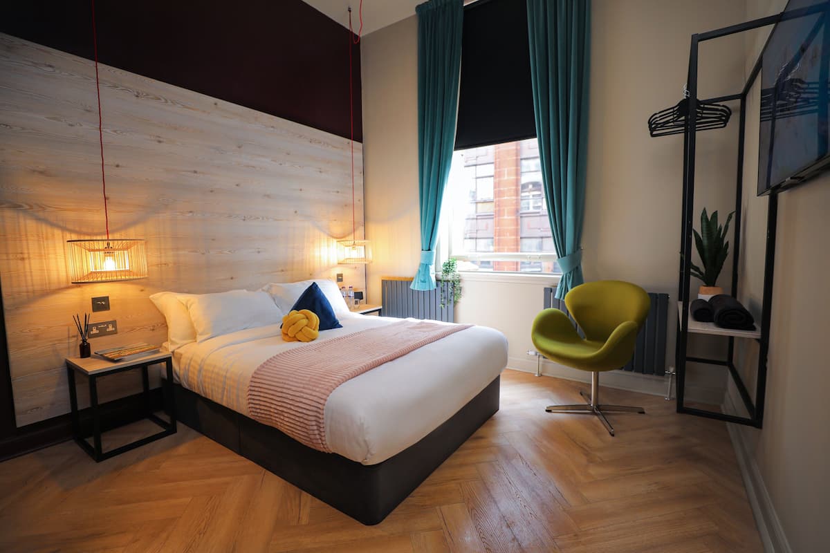 Ein modernes Schlafzimmer mit einem großen Bett, Nachttischlampen, einem grünen Sessel und Blick auf die Stadt durch ein Fenster.