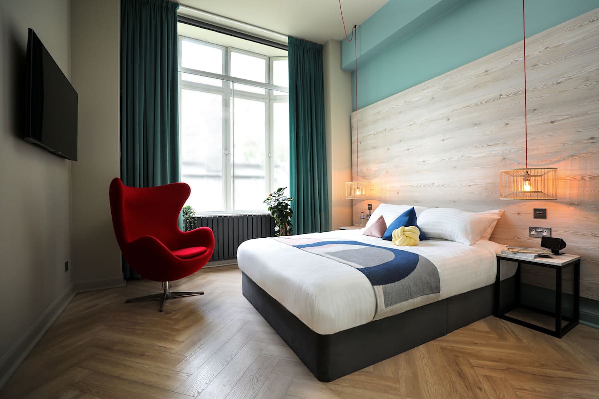 Ein modernes Hotelzimmer mit einem großen Bett, einem roten Sessel, einem Fernseher und einem Fenster mit Vorhängen.