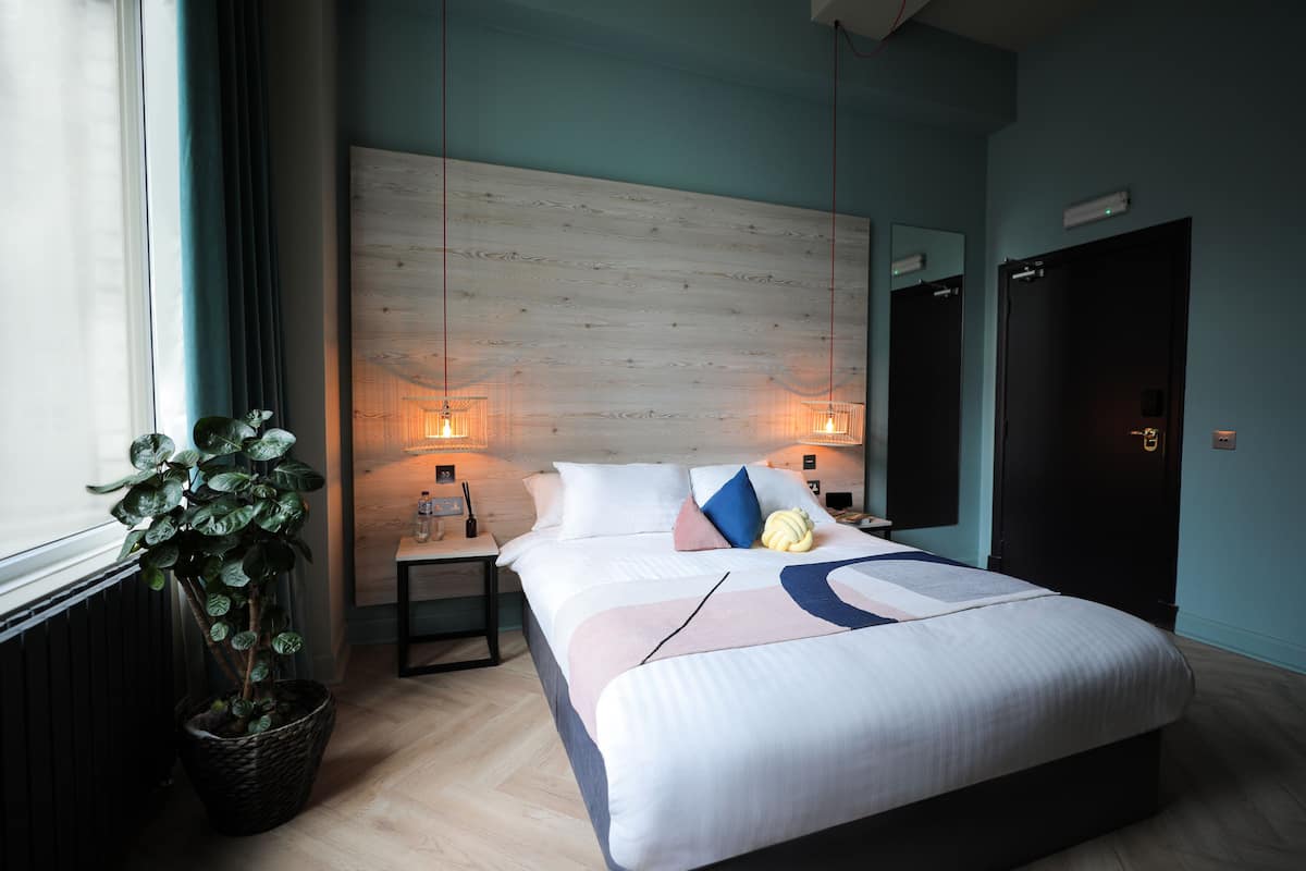 Ein modernes Hotelzimmer mit einem großen Bett, Nachttischlampen, einer Topfpflanze und einem Holz-Kopfstück.