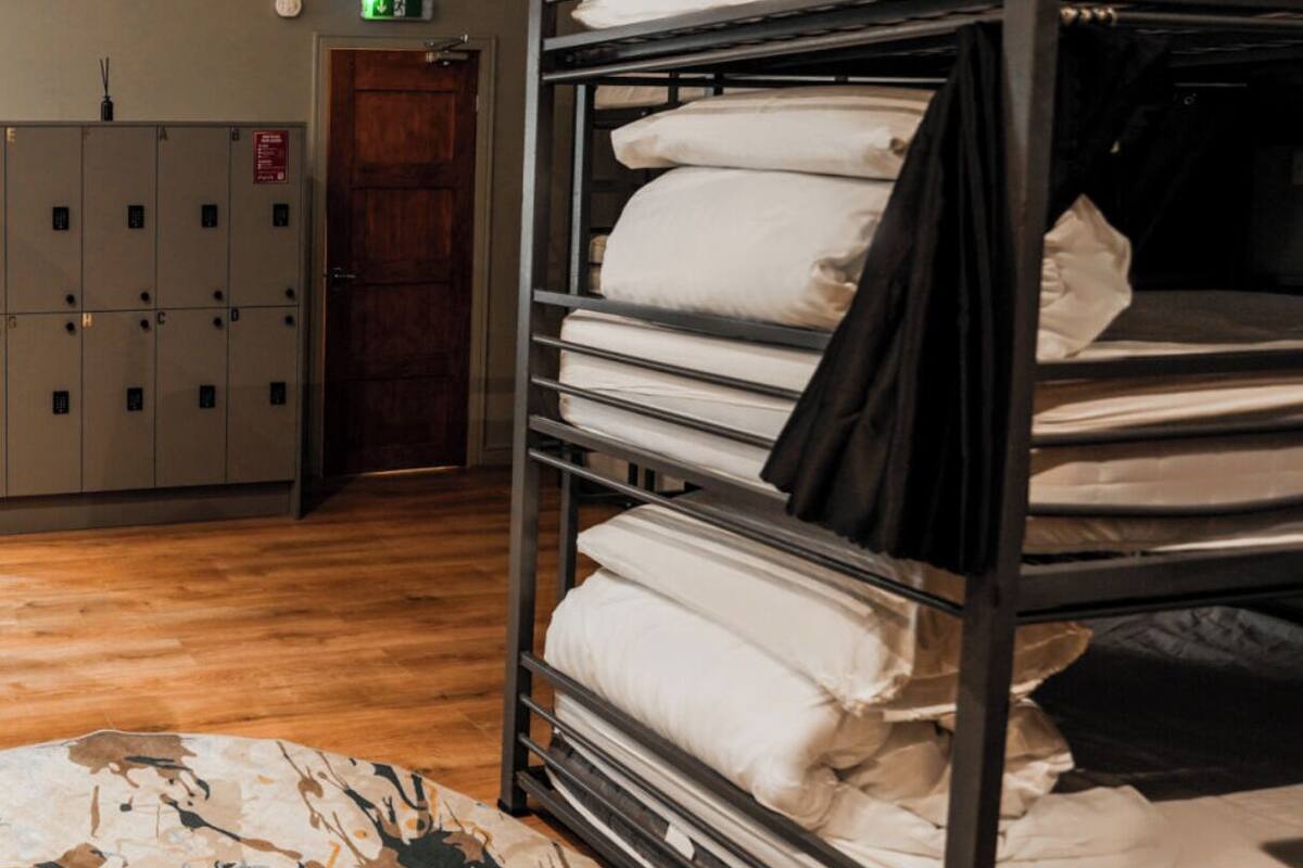 Ein Stockbett mit weißer Bettwäsche, ein Holzboden und ein gemustertes Teppich. Im Hintergrund sind Schließfächer und eine Tür zu sehen.