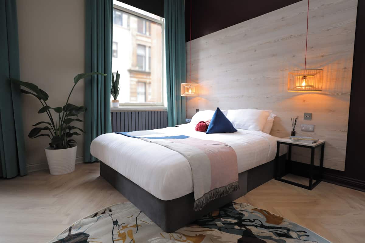 Ein modernes Schlafzimmer mit einem Bett, Nachttischlampen, einer Topfpflanze und Blick auf die Stadt durch ein Fenster.