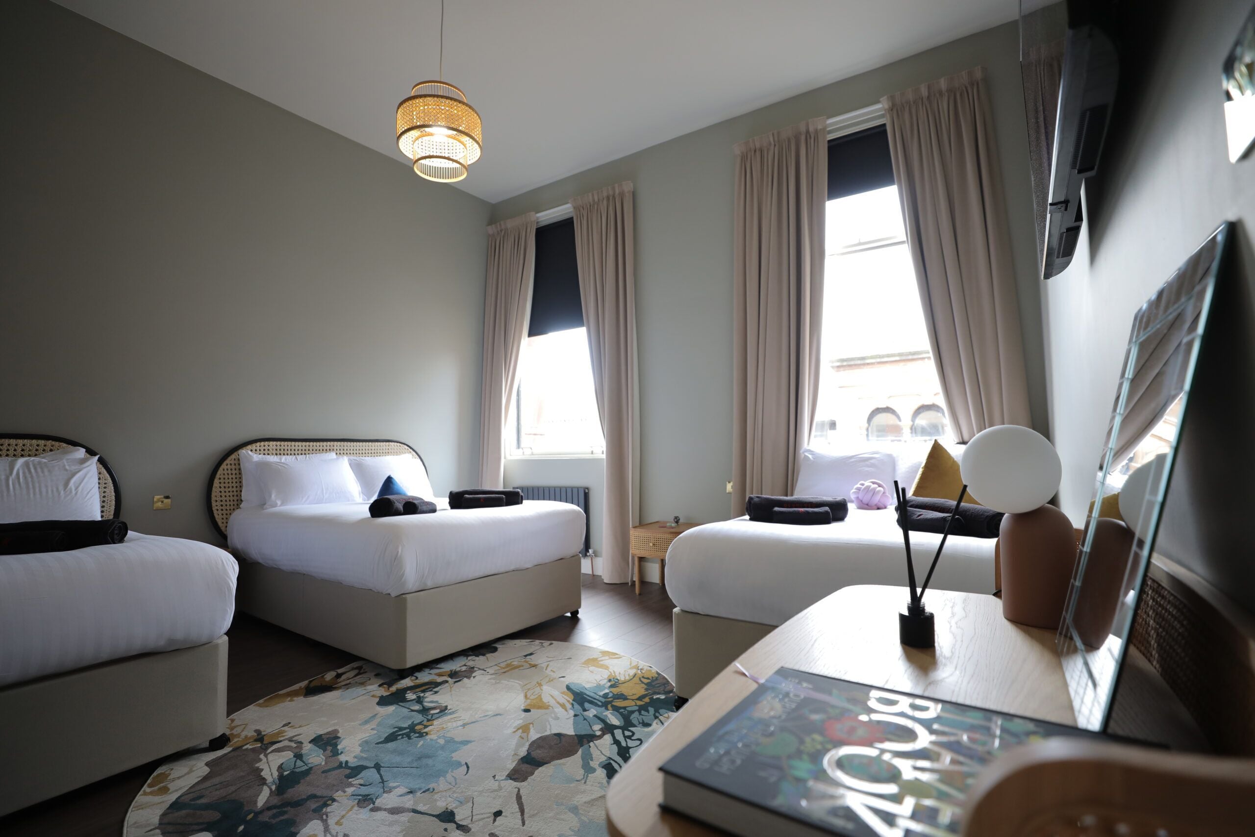 A hotel room with two beds, a small table, a lamp, and a large window with curtains.