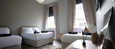 A hotel room with two beds, a small table, a lamp, and a large window with curtains.