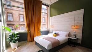 A modern bedroom with a large bed, a potted plant, a view of a building through a window, and a wooden headboard. - Revolver Glasgow (Glasgow)