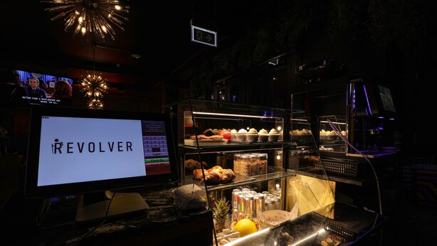 A bar area with a digital screen displaying "REVOLVER," a glass display case with pastries and drinks, and a bar counter with a cash register. - Revolver Glasgow (Glasgow)
