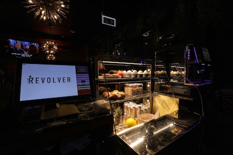 A bar area with a digital screen displaying "REVOLVER," a glass display case with pastries and drinks, and a bar counter with a cash register.