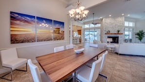 A spacious dining area with a wooden table and white chairs, a chandelier, and a large painting of a sunset on the wall.
