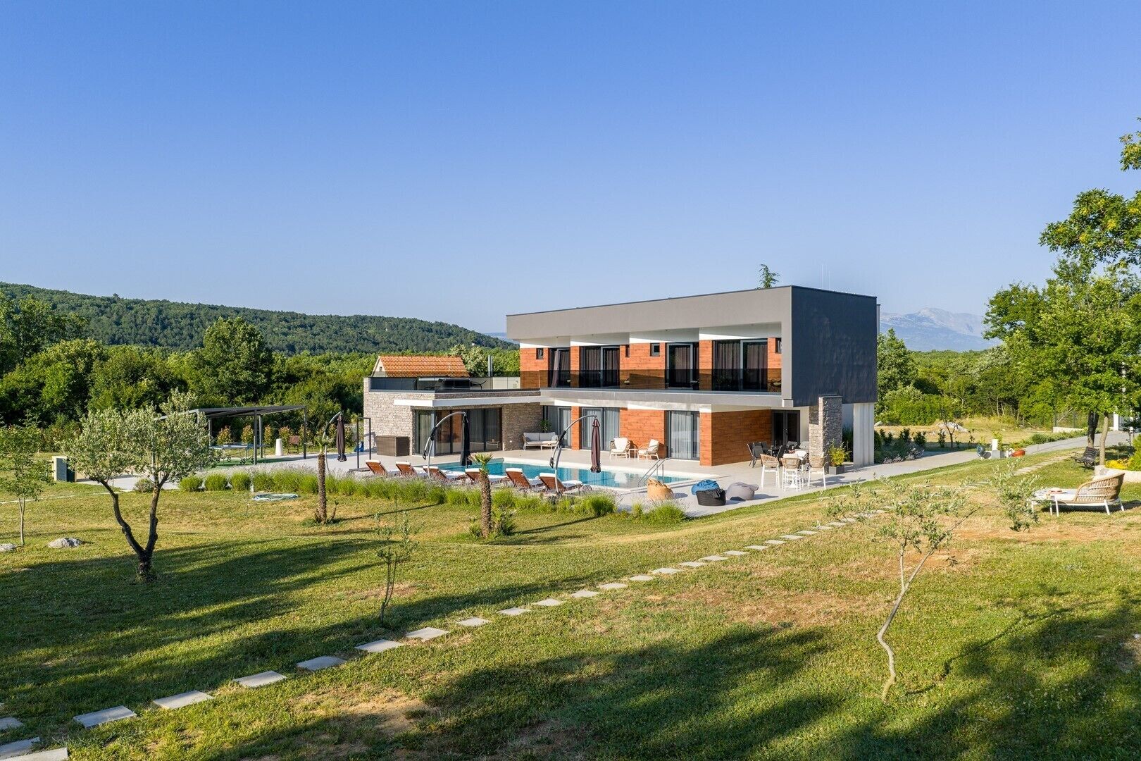 A modern two-story building with a swimming pool, outdoor seating, and a view of the mountains.