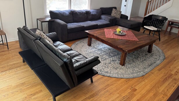 Una sala de estar con un sofá modular gris oscuro, una mesa de centro de madera con un objeto decorativo, un tapete estampado y una escalera al fondo.