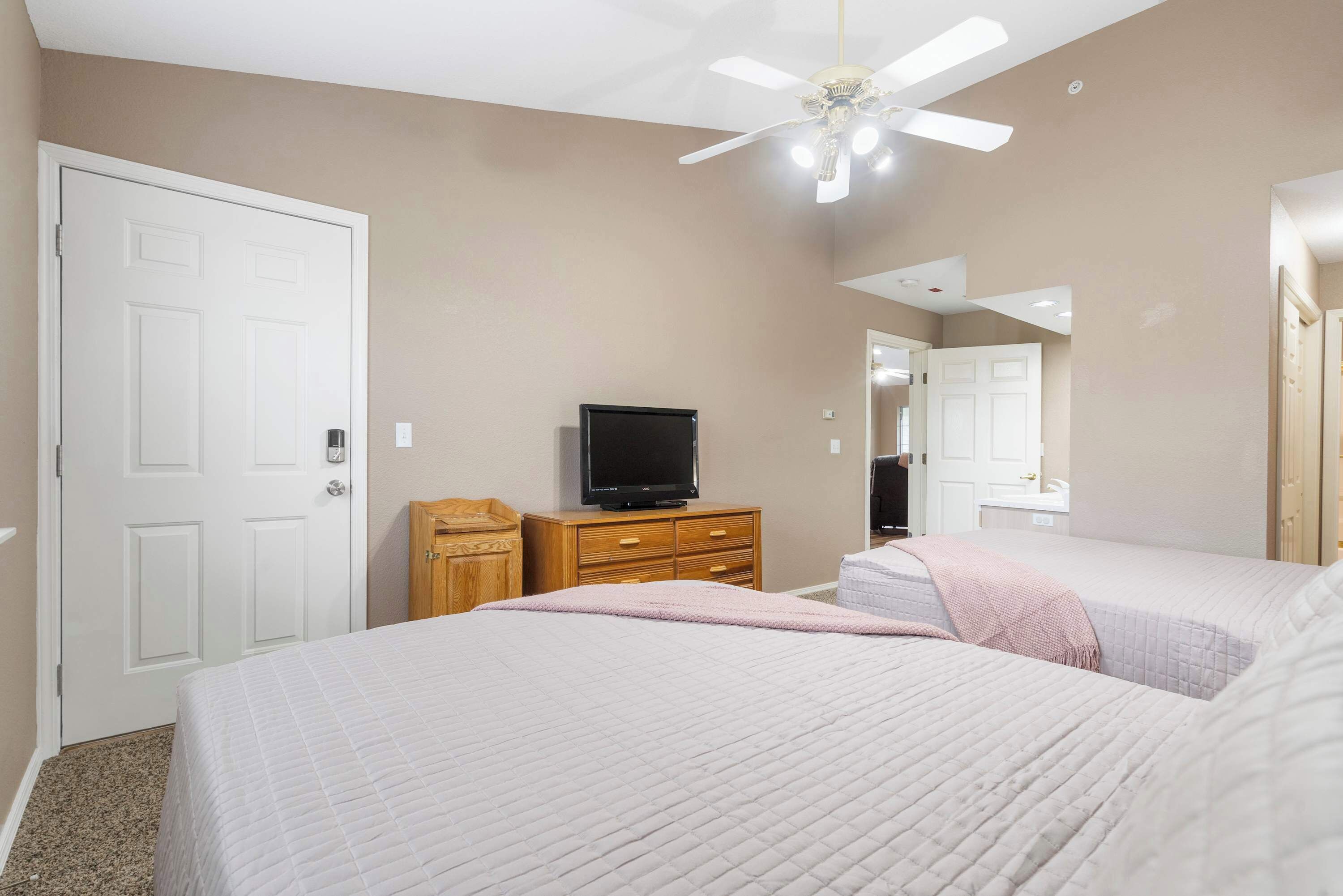 A bedroom with two beds, a television on a wooden dresser, a ceiling fan, and a white door.