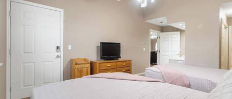 A bedroom with two beds, a television on a wooden dresser, a ceiling fan, and a white door.