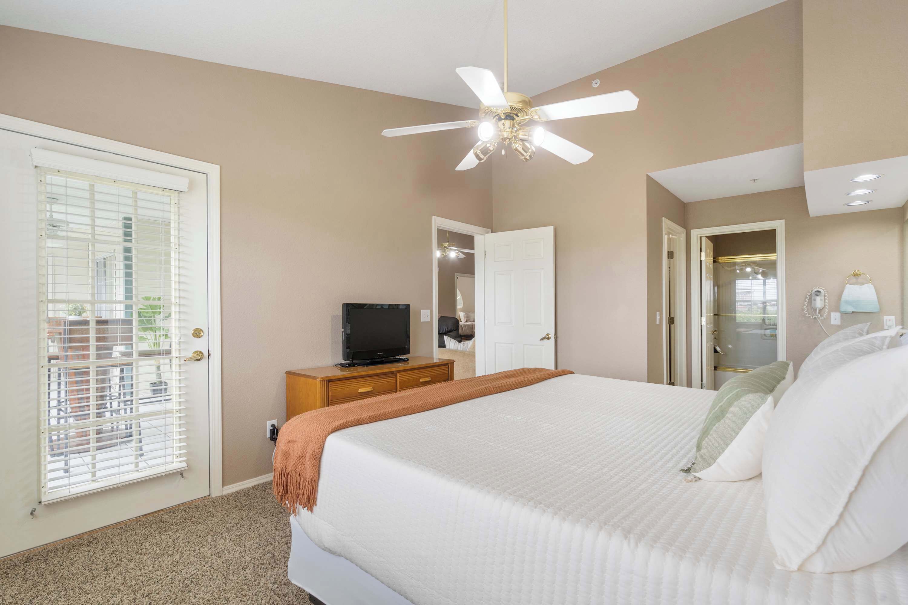 Une chambre à coucher avec un lit, un ventilateur de plafond, un téléviseur posé sur une commode en bois et une porte donnant sur une autre pièce.