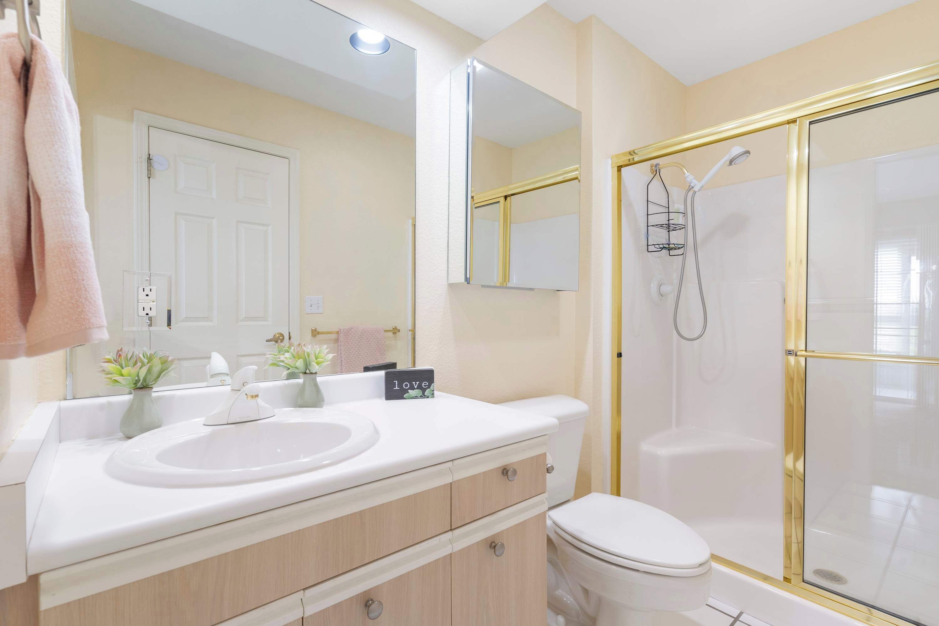 Une salle de bain moderne avec un lavabo, un meuble-lavabo et des toilettes, le tout de couleur blanche. Elle comprend une douche à l’italienne avec un encadrement doré et un grand miroir au-dessus du meuble-lavabo.