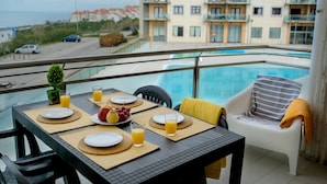 Une table à manger dressée avec des assiettes, des verres et un saladier de fruits, donnant sur une piscine et un immeuble doté de balcons.