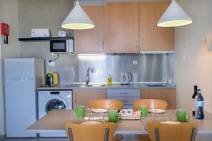 A compact kitchen with a washing machine, microwave, and a sink. There is a dining table with chairs and a basket of snacks.