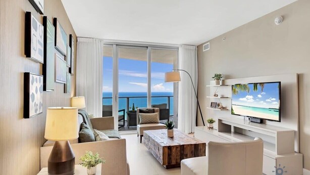 A modern living room with a large TV, comfortable seating, a coffee table, and a balcony with a view of the sea. - Ocean View Balcony|Beachfront Resort |Amazing Pool (Hollywood)