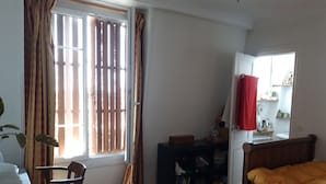A bedroom with a wooden floor, a bed with an orange blanket, a desk with a chair, a wardrobe, and a window with curtains.