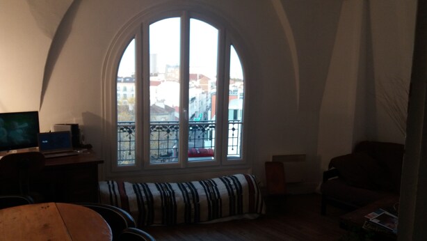 A room with a large arched window, a wooden bench, a television, and a small table with speakers.