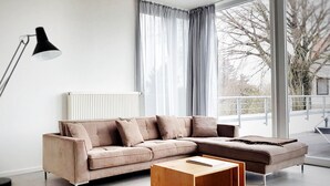 Een moderne woonkamer met een hoekbank, een houten bijzettafeltje, een vloerlamp en een groot raam met gordijnen.