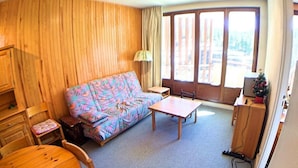 Una habitación con pared de madera, un sofá colorido, una mesita, una silla y un ventanal con vistas a árboles.