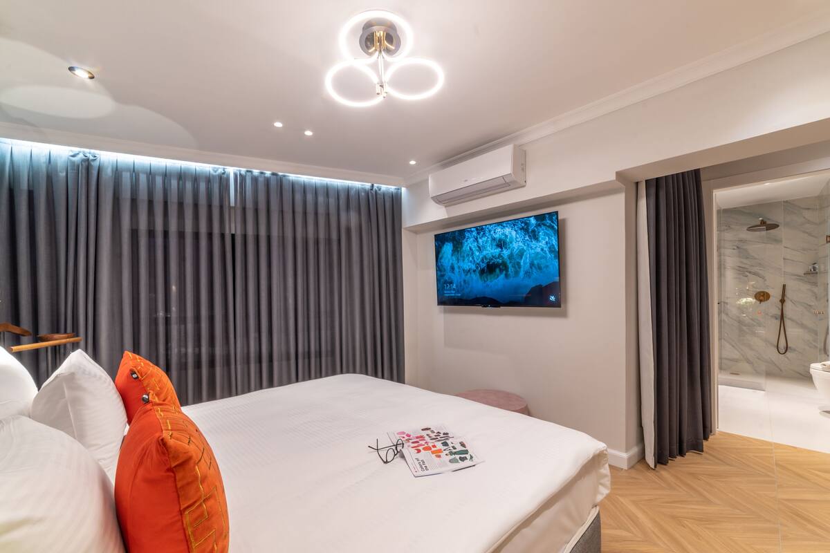 A modern hotel room with a large bed, a flat-screen TV mounted on the wall, a shower area with marble walls, and a wooden floor.