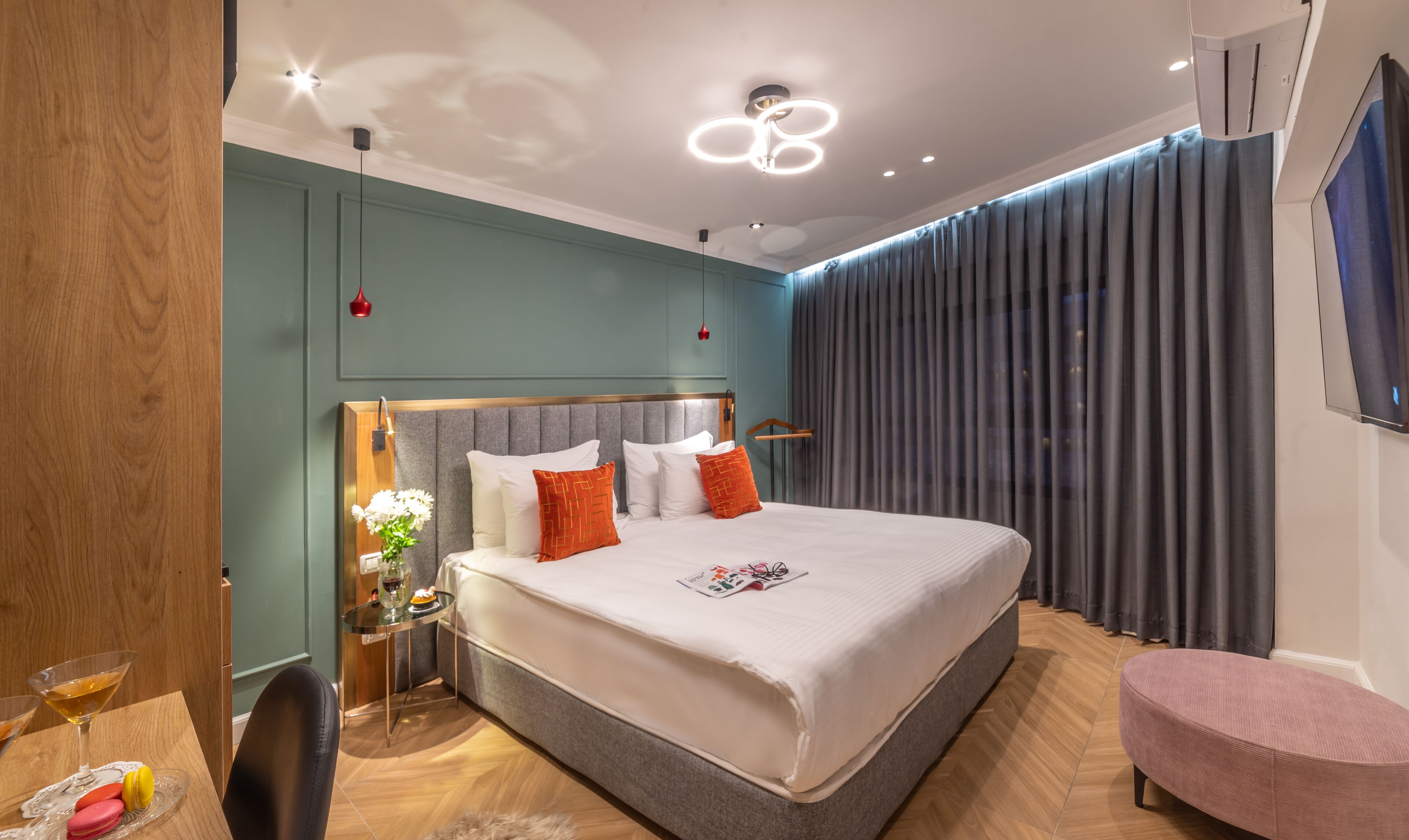 A modern hotel room with a large bed, a bedside table with a vase of flowers, a small round table with a drink and snacks, and a large window with curtains.