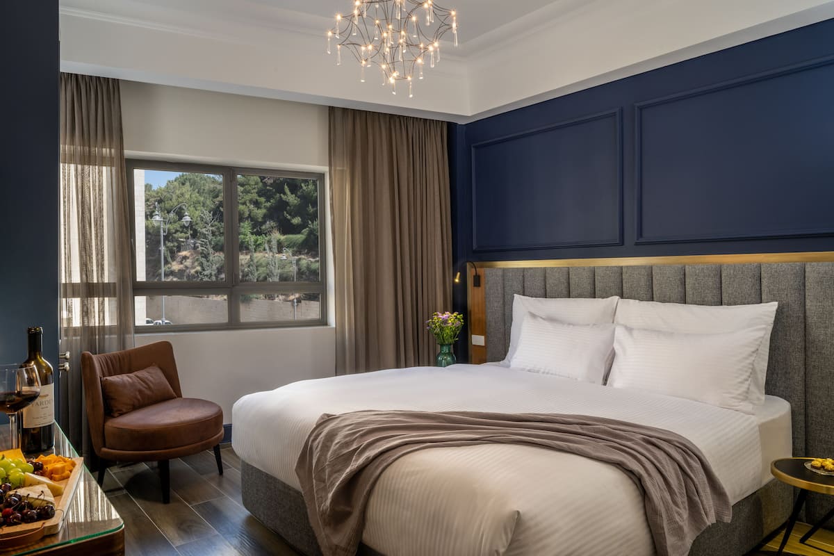 A modern hotel room with a large bed, a sitting area with a chair, a small table with a bottle of wine, and a view of the outdoors.