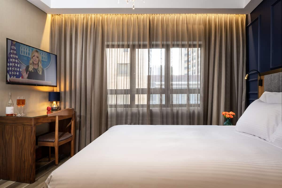 A hotel room with a large bed, a desk with a chair, a television displaying a news anchor, and a window with curtains.