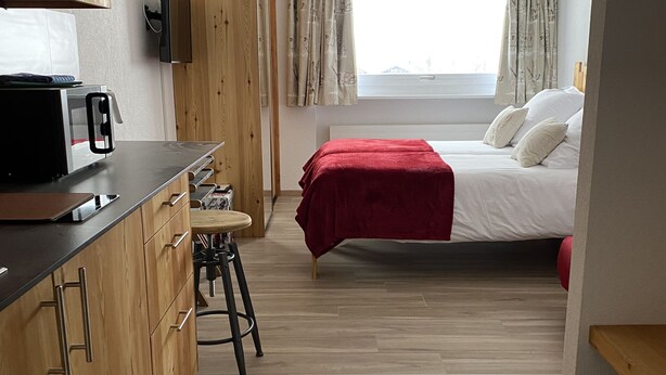 A compact room with a kitchenette, a bed with a red blanket, and a window with curtains.