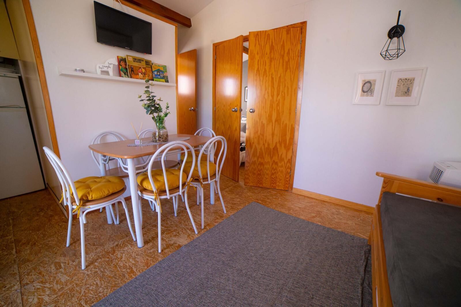 A compact living space with a dining area featuring a table and chairs, a television mounted on the wall, and a wooden door leading to another room.