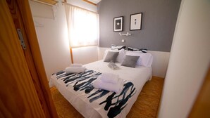 Un dormitorio con cama, almohadas, una manta y una toalla. Hay una ventana con cortinas, un teléfono de pared y dos cuadros enmarcados.