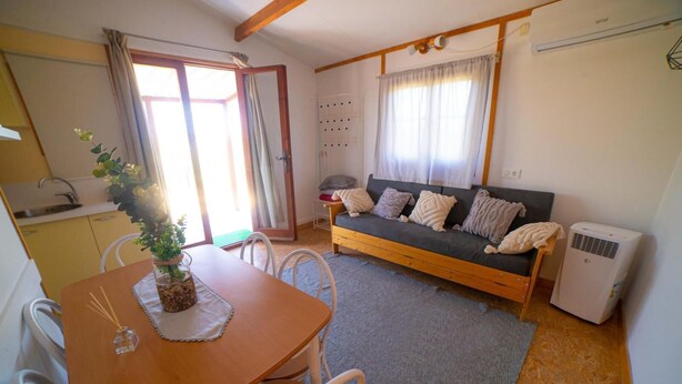 Una sala de estar acogedora con un sofá, una mesa de comedor con sillas, una zona de cocina y un gran ventanal con cortinas.