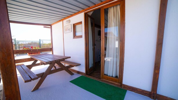 Eine überdachte Terrasse mit einem Holztisch und Bänken, einer Schiebetür und einer grünen Fußmatte im Eingangsbereich.