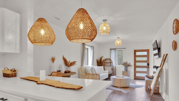 A spacious living area with a white sofa, a coffee table, and a large window. There are three hanging wicker lamps and a flat-screen TV mounted on the wall.