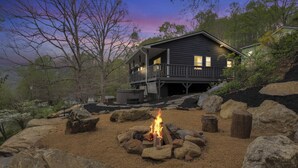 A cabin with a fire pit surrounded by rocks, set in a natural landscape with trees and a house in the background.