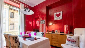 A dining area with a table set for two, chairs with floral upholstery, a red tiled wall, and a white cushioned chair with a car pillow. - Billie's Flat RED - art & design apt in Verona historic centre (Verona)